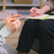 A close up of two people hands. The people are sitting across from each other. One person is writing in a notebook.
