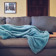 Someone laying on a grey couch with a blue blanket draped over their head and torso.