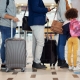 People standing in line at an airport check-in counter with luggage and travel bags, including a small child wearing a pink backpack.